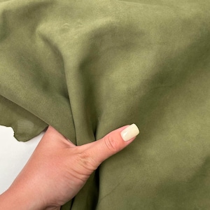 May include: Close-up of olive green fabric. The fabric is soft and appears to be suede or a similar material. The texture is smooth with subtle variations in color. The fabric is being held by a hand with a light-colored nail polish.