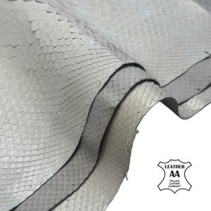 May include: Close-up of a stack of light gray snake skin leather. The leather has a scale pattern and is folded over itself. The image includes the logo of the Italian Leather Company.