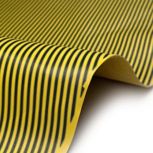 Puede incluir: Primer plano de una lámina de material amarillo con rayas verticales negras. El material está ligeramente curvado, revelando una vista de la parte inferior. Las rayas están espaciadas uniformemente y recorren la longitud del material.