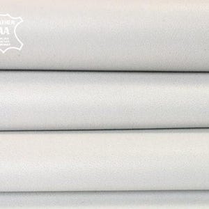May include: A stack of folded white leather sheets with a smooth, matte texture. In the top left corner, a white emblem features the text LEATHER AA ITALIAN LEATHER COMPANY inside a stylized hide shape, indicating the material origin.