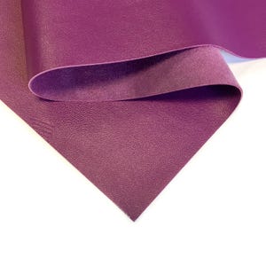May include: A sheet of deep purple faux leather with a textured surface. The material is folded over, revealing a lighter, suede-like underside. The color is a rich, saturated hue, suitable for various crafting projects.