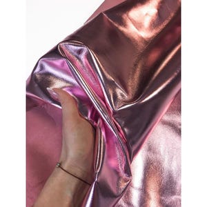 Può includere: Primo piano di un tessuto rosa metallizzato lucido. Il tessuto è piegato e ha una superficie riflettente, con luci e ombre che creano un senso di profondità. Il materiale sembra liscio e lussuoso.