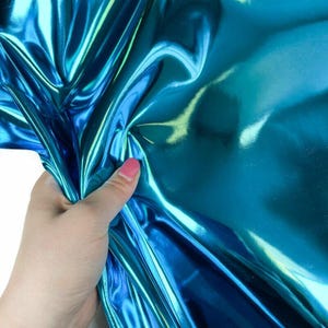 May include: A hand holding a piece of shiny, iridescent teal fabric. The fabric has a metallic sheen and reflects light, creating highlights and shadows. The material appears to be soft and pliable.