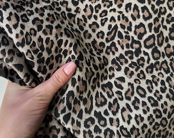 May include: Close-up of a fabric with a leopard print pattern. The fabric features a tan background with dark brown and black spots. The fabric is being held, showing its texture and drape.