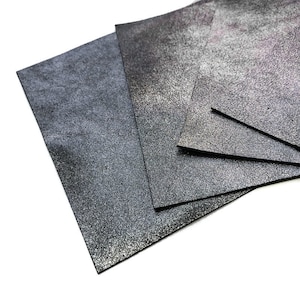 May include: Several sheets of textured, dark gray leather are arranged on a white surface. The leather has a speckled, metallic appearance, with variations in tone from charcoal to silver. The edges of the leather are cleanly cut, and the overall presentation is neat.