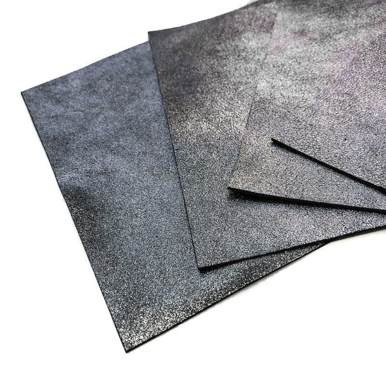 May include: Several sheets of textured, dark gray leather are arranged on a white surface. The leather has a speckled, metallic appearance, with variations in tone from charcoal to silver. The edges of the leather are cleanly cut, and the overall presentation is neat.