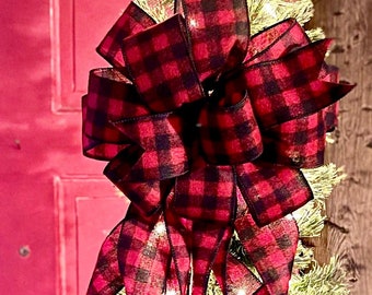 Pode incluir: Um laço de fita vermelha e preta xadrez está amarrado no topo de uma árvore de Natal artificial verde. A árvore está decorada com luzes brancas.