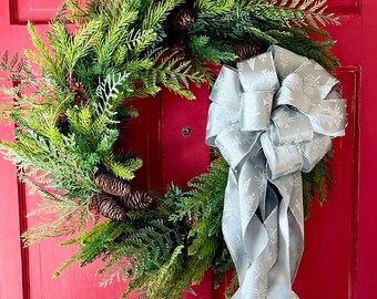 Pode incluir: Uma grinalda de pinho verde com pinhas marrons e um laço prateado com um padrão de floco de neve pendura em uma porta vermelha.