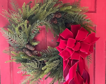 Pode incluir: Uma grinalda verde de sempre-verde com pinhas pendura em uma porta vermelha com um grande laço de veludo vermelho brilhante.