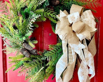 Pode incluir: Uma guirlanda de Natal verde com agulhas de pinho e pinhas, decorada com um grande laço dourado e branco com padrões de flocos de neve.