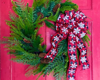 Pode incluir: Uma coroa de Natal feita de ramos de pinho verdes com pinhas e um laço xadrez vermelho e preto com flocos de neve brancos.