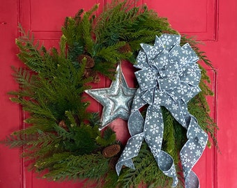 Pode incluir: Uma grinalda verde de sempre-verde com uma estrela prateada e um laço cinza com estampa de floco de neve branco. A grinalda está pendurada em uma porta vermelha.
