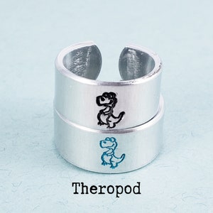 Puede incluir: Dos anillos ajustables de color plateado apilados uno encima del otro. Cada anillo tiene una ilustraci&oacute;n de dinosaurio de dibujos animados. El anillo superior tiene un dinosaurio negro y el inferior uno azul. La palabra "Theropod" est&aacute; impresa debajo.
