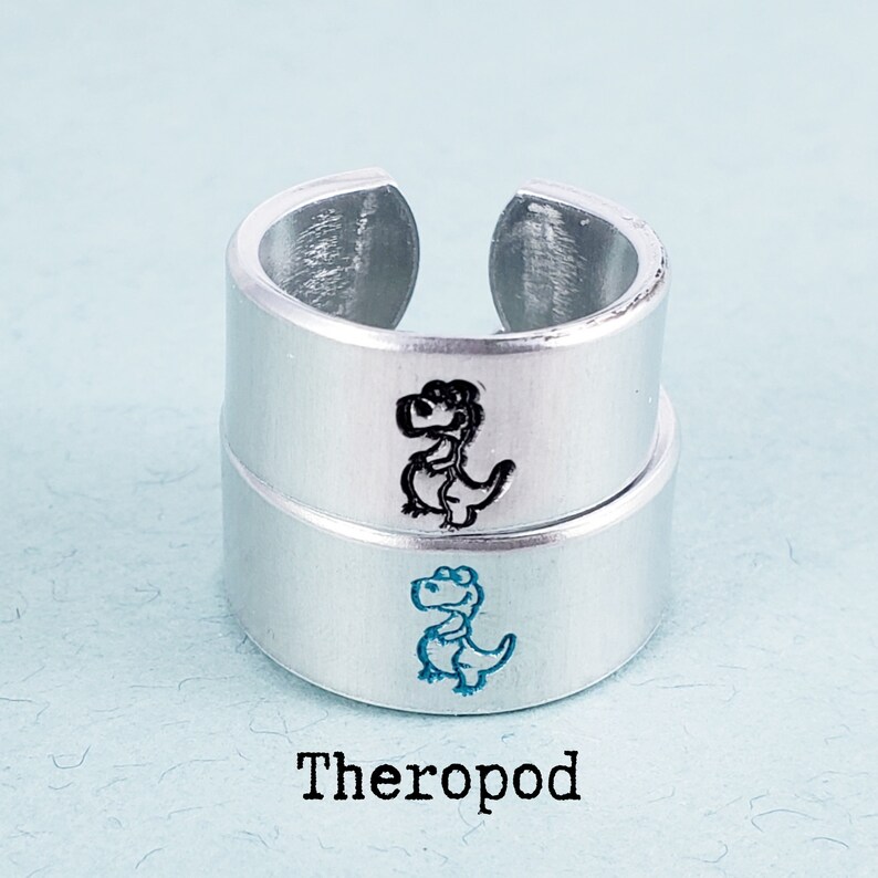 Puede incluir: Dos anillos ajustables de color plateado apilados uno encima del otro. Cada anillo tiene una ilustraci&oacute;n de dinosaurio de dibujos animados. El anillo superior tiene un dinosaurio negro y el inferior uno azul. La palabra "Theropod" est&aacute; impresa debajo.