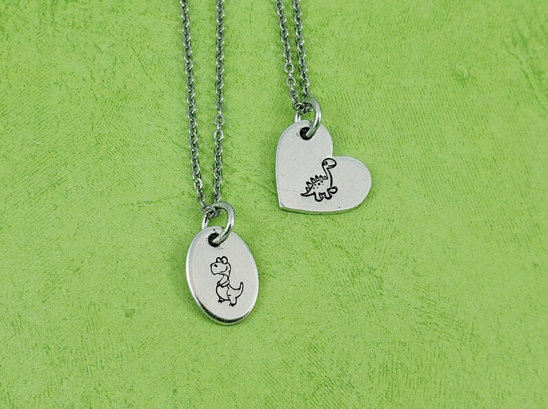 Puede incluir: Dos collares con colgantes de dinosaurio plateados. Un colgante es ovalado, el otro en forma de coraz&oacute;n. Cada colgante presenta una ilustraci&oacute;n de dinosaurio negro y cuelga de una delicada cadena. El fondo es verde texturizado.