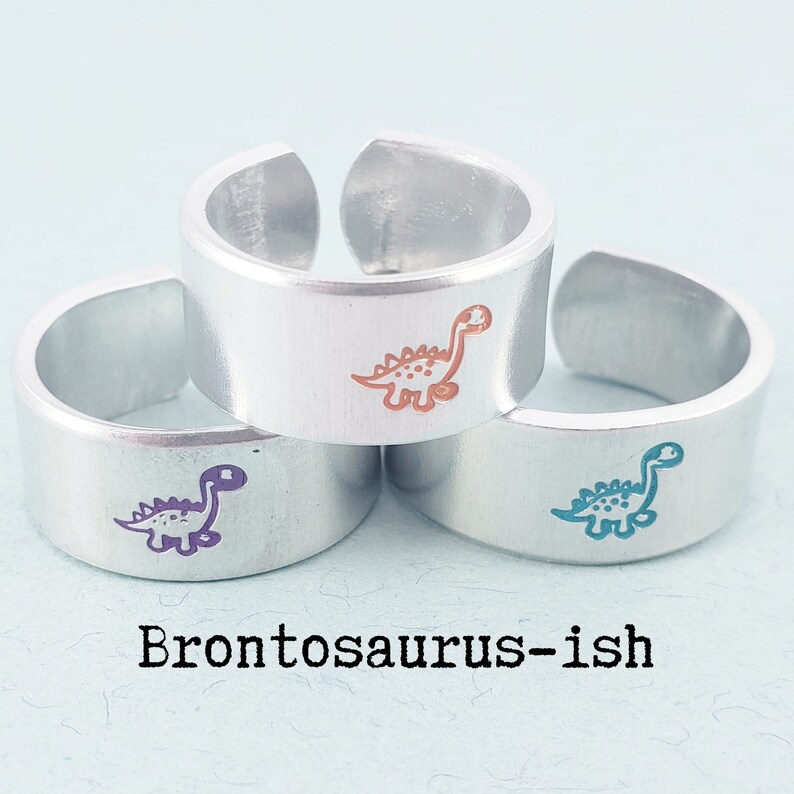Puede incluir: Tres anillos ajustables de plata con ilustraciones de dinosaurios en morado, naranja y turquesa. Los anillos est&aacute;n estampados con la palabra "Brontosaurus-ish".