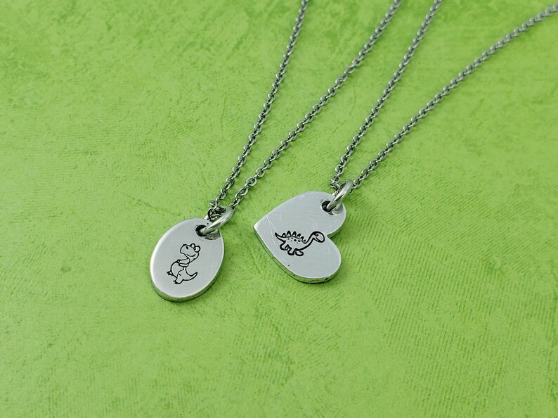 Puede incluir: Dos collares plateados con colgantes de dinosaurios. Un colgante es ovalado, el otro en forma de coraz&oacute;n. Ambos colgantes tienen una peque&ntilde;a ilustraci&oacute;n de dinosaurio negro. Sobre fondo verde.