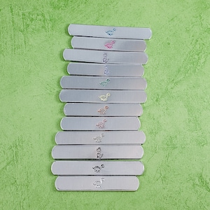 Puede incluir: Una colecci&oacute;n de barras de metal plateado, cada una adornada con un dise&ntilde;o de dinosaurio de diferente color. Los dinosaurios son de varios colores, incluyendo azul, rosa, verde, amarillo y naranja. Las barras est&aacute;n dispuestas sobre una superficie verde texturizada.