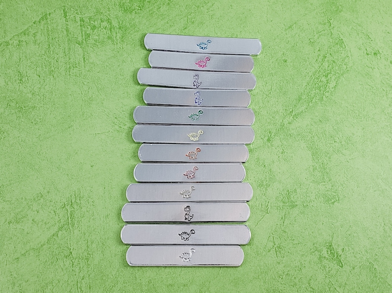 Puede incluir: Una colecci&oacute;n de barras de metal plateado, cada una adornada con un dise&ntilde;o de dinosaurio de diferente color. Los dinosaurios son de varios colores, incluyendo azul, rosa, verde, amarillo y naranja. Las barras est&aacute;n dispuestas sobre una superficie verde texturizada.