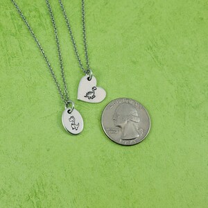 Puede incluir: Tres collares plateados con colgantes de dinosaurios. Un colgante tiene forma de coraz&oacute;n y el otro es ovalado. Una moneda de veinticinco centavos estadounidense est&aacute; junto a los collares para comparar el tama&ntilde;o. El fondo es verde liso.