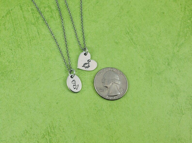 Puede incluir: Tres collares plateados con colgantes de dinosaurios. Un colgante tiene forma de coraz&oacute;n y el otro es ovalado. Una moneda de veinticinco centavos estadounidense est&aacute; junto a los collares para comparar el tama&ntilde;o. El fondo es verde liso.