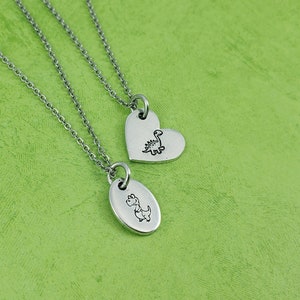 Puede incluir: Dos collares plateados con colgantes. Uno tiene un colgante en forma de coraz&oacute;n, el otro un colgante ovalado. Cada colgante presenta una peque&ntilde;a ilustraci&oacute;n de dinosaurio negro. Las cadenas son delicadas y el fondo es verde.