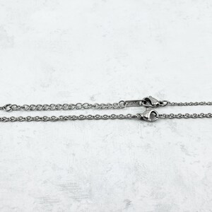 Puede incluir: Dos collares plateados con diferentes estilos de cadena. Uno tiene una cadena fina, el otro un dise&ntilde;o ligeramente m&aacute;s grueso. Ambos collares tienen un cierre y se muestran sobre un fondo claro y texturizado.
