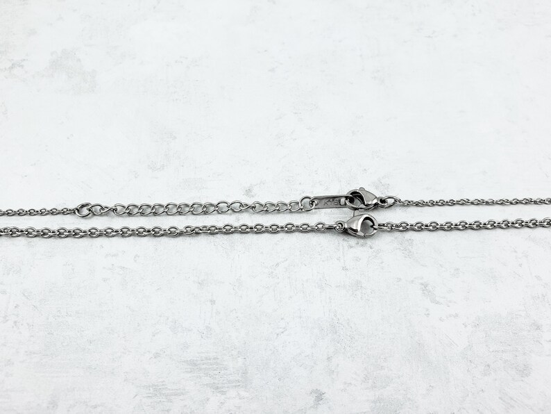 Puede incluir: Dos collares plateados con diferentes estilos de cadena. Uno tiene una cadena fina, el otro un dise&ntilde;o ligeramente m&aacute;s grueso. Ambos collares tienen un cierre y se muestran sobre un fondo claro y texturizado.
