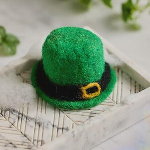 May include: A small, green felt hat with a black band and a gold buckle. The hat is a miniature version of a traditional Irish leprechaun hat, often worn for St. Patrick's Day. It sits on a white marble tray.
