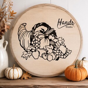 May include: A round light-colored wooden sign featuring a black graphic of a cornucopia filled with harvest produce including pumpkins, grapes, and corn. The text Handr Studio is printed in black in the upper right corner of the wooden surface.