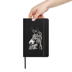 May include: Black notebook with a white illustration of a donkey's head and the name "Olivia" printed on the cover.