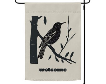 May include: A beige garden flag with a black silhouette of a bird perched on a branch. The text "welcome" is printed in black at the bottom of the flag.
