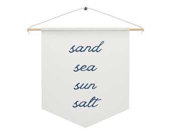 May include: A white banner with the words "sand, sea, sun, salt" in a dark blue script. The banner is suspended from a wooden rod with a string and a small metal fastener.
