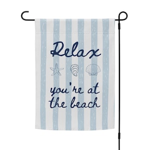 May include: A garden flag with blue and white vertical stripes. The flag has the words "Relax you're at the beach" in dark blue script, along with illustrations of a starfish, oyster, and seashell.