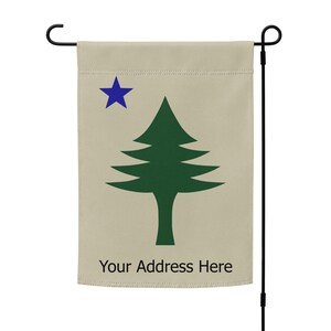 Op de afbeelding: Een beige tuinvaandel met een donkergroene dennenboom en een blauwe ster. De tekst "Your Address Here" staat onderaan afgedrukt. De vlag hangt aan een zwarte metalen paal.