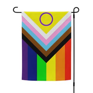 Puede incluir: Una bandera de jardín con un diseño de bandera del orgullo arcoíris, que presenta un círculo morado con un centro amarillo en la sección superior. La bandera está unida a un poste de metal negro.