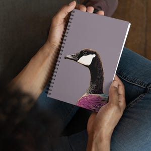 May include: A spiral bound notebook with a purple cover featuring a colorful illustration of a goose with a white head and neck. The goose is facing left and has a pink, purple, and blue body.