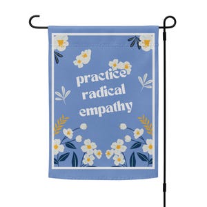 May include: A blue garden flag with white flowers and the text "practice radical empathy" in white.