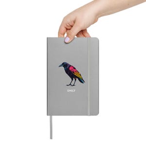 May include: A gray notebook with a colorful illustration of a crow. The crow is depicted with a black head, a pink chest, and yellow and blue wings. The name "EMILY" is printed below the crow.