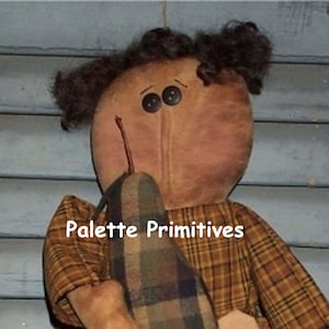 May include: A handmade rag doll with brown fabric skin, black button eyes, and curly brown hair. The doll is wearing a plaid dress with brown and orange squares. The doll is holding a brown and green plaid fabric in its hands. The text "Palette Primitives" is visible on the doll's dress.