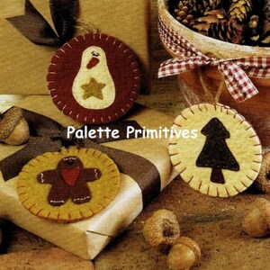 Peut inclure: Trois décorations de Noël en feutre avec un bonhomme en pain d'épices, un sapin de Noël et un pingouin. Les décorations sont beige, marron et rouge avec des coutures jaunes. Le texte "Palette Primitives" est visible sur l'image.