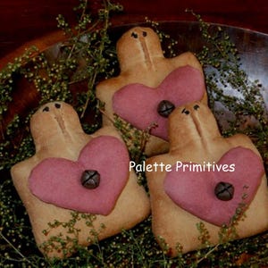 Könnte beinhalten: Drei herzförmige primitive Volkskunst-Ornamente mit rosa Herzen und braunem Stoff. Die Ornamente liegen in einer Holzschale mit grünem Laub.