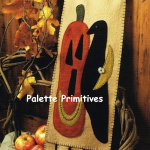 May include: A felt wall hanging with a black crow and a red pumpkin with a yellow stem. The background is beige and the hanging has a brown fringe. The text "Palette Primitives" is printed on the hanging.