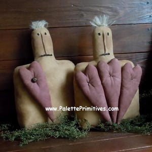 May include: Two primitive-style dolls with tan bodies and mauve heart-shaped hands. Each doll has a tuft of white hair and simple facial features. One doll holds a single heart, the other holds three. The website www.PalettePrimitives.com is visible.