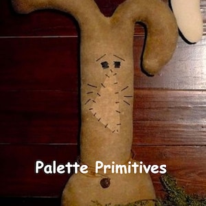 May include: A brown felt bunny rabbit with a stitched face and a brown felt tail. The bunny is standing on a bed of green foliage. The text "Palette Primitives" is visible below the bunny.