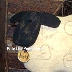 Puede incluir: Una oveja blanca y negra con la cara negra y una etiqueta marrón que dice "Baa" colgando de su cuello. La oveja está sentada sobre una superficie de madera.