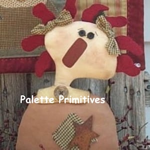 Könnte beinhalten: Eine handgemachte Stoffpuppe mit roten Haaren, einem braunen und weißen karierten Kleid und einem sternförmigen Patch. Die Puppe steht auf einer Holzoberfläche und hat den Text "Palette Primitives" auf der Vorderseite des Kleides.