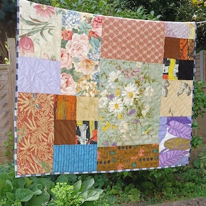 May include: A patchwork quilt with a variety of floral and patterned fabric squares. The quilt is displayed outdoors, hanging on a washing line. The colour scheme includes pink, blue, green, and brown. The quilt is a handmade item.