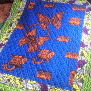 May include: A quilted textile featuring a vibrant blue central panel with orange butterfly and floral designs. The quilt is bordered with green, yellow, purple, and white floral patterns, creating a colorful and textured piece.