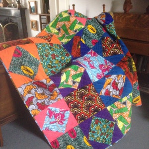 May include: A patchwork quilt featuring a vibrant mix of geometric patterns in orange, blue, green, and purple. The quilt is draped over a wooden headboard, displaying its intricate design and craftsmanship. The quilt's measurements are not visible.